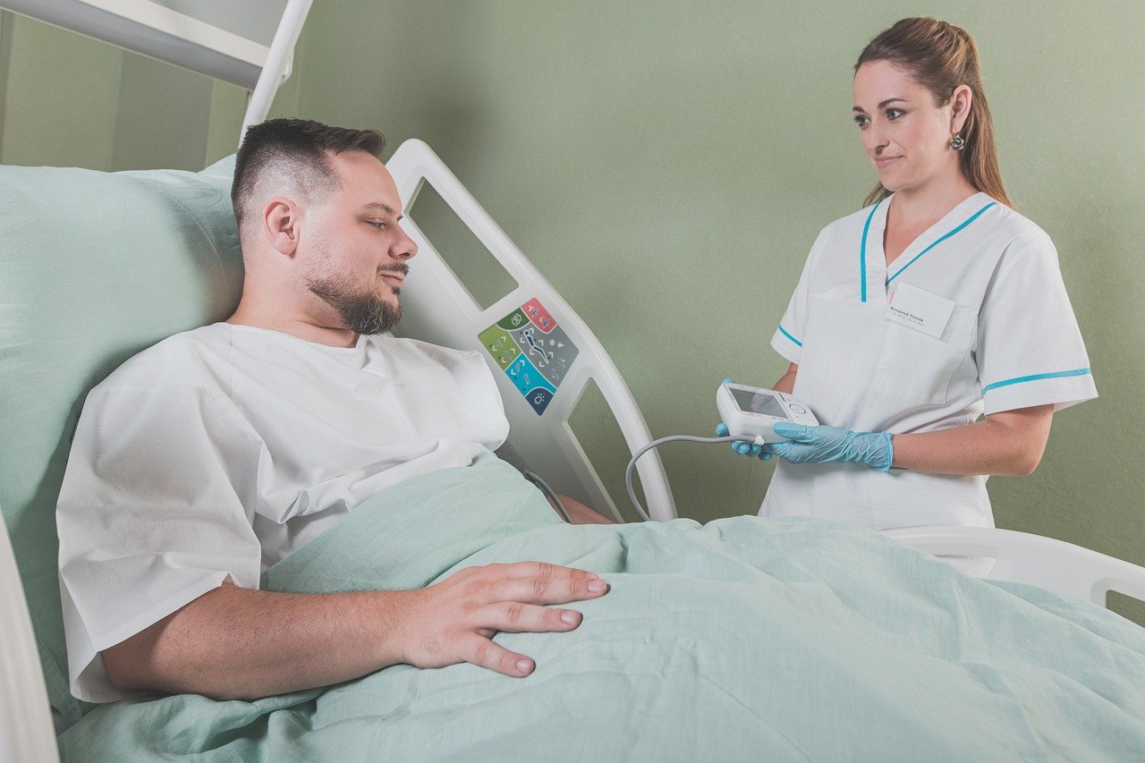 Ein Patient liegt in einem Bett, dessen Kopfteil hochgefahren wurde, daneben steht eine Mitarbeiterin und misst seinen Blutdruck.